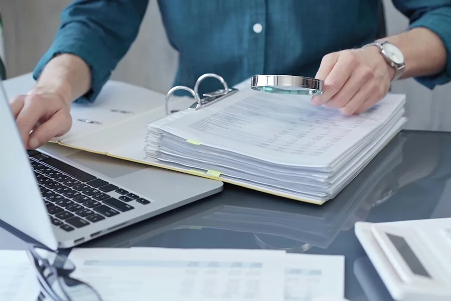 Personne qui lit des lignes comptables sur un document papier dans un classeur à l'aide d'une loupe